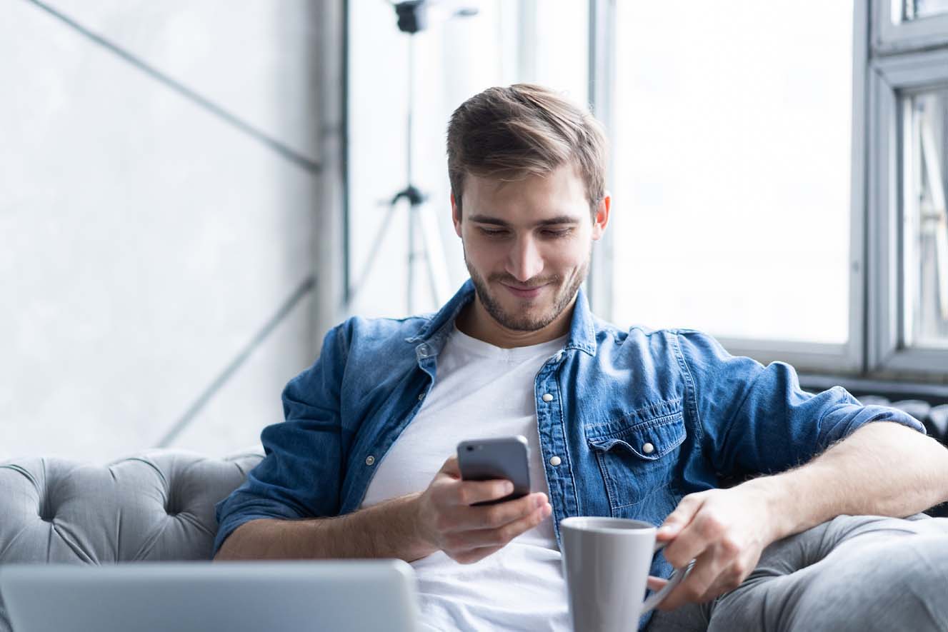 A young man in his living room drinking coffee and using his smartphone